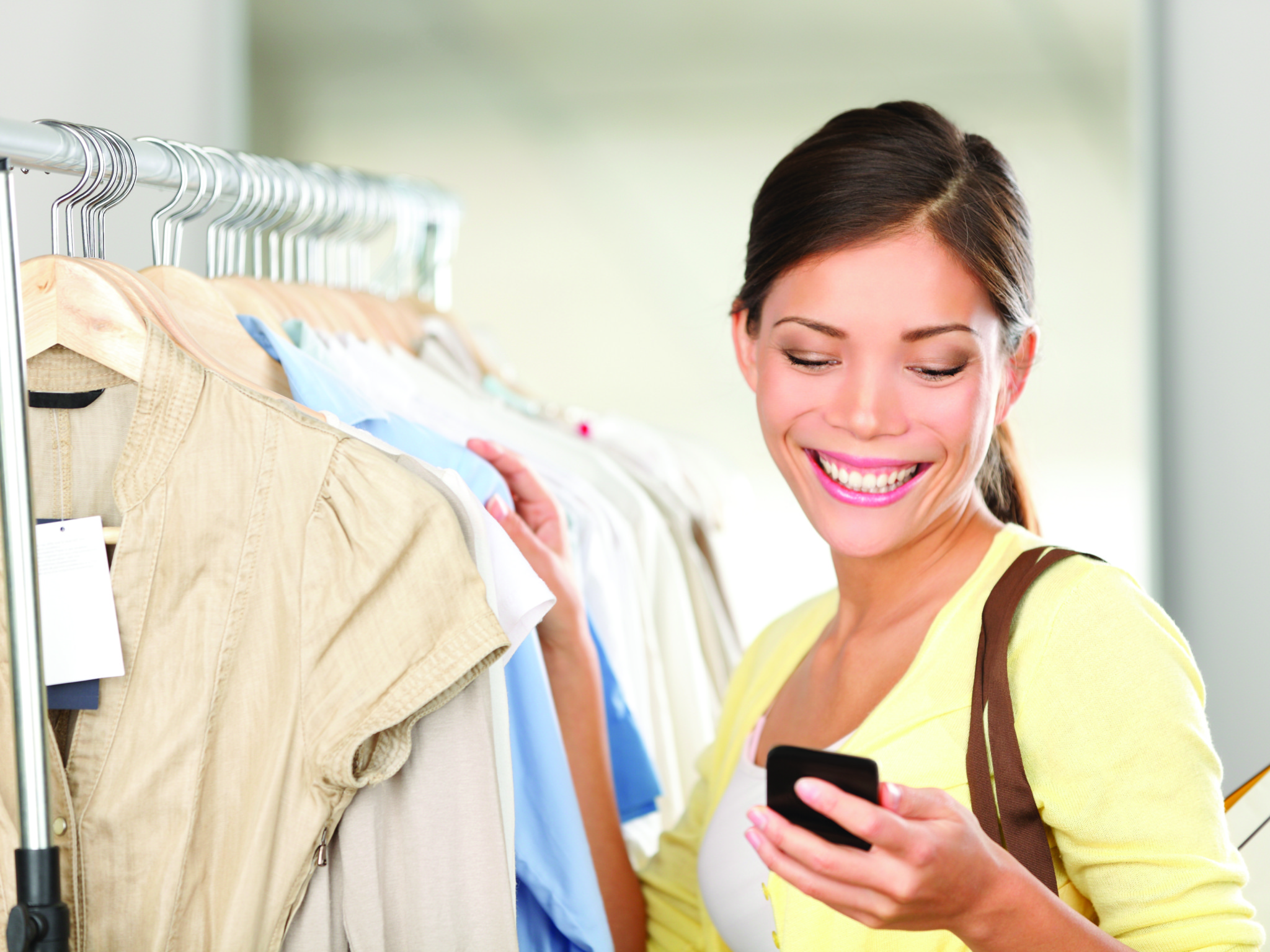 Modern woman shopping looking at smartphone texting or talking smiling happy in clothes store. Beautiful young mixed race Asian / Caucasian young woman shopper.